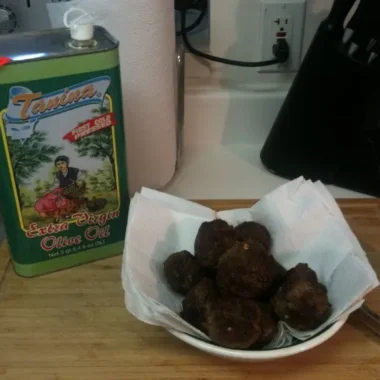 Bowl of homemade Italian meatballs resting on paper towels next to a tin of extra virgin olive oil on a kitchen counter.
