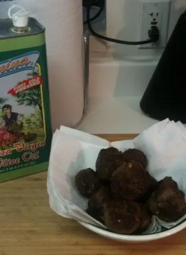Bowl of homemade Italian meatballs resting on paper towels next to a tin of extra virgin olive oil on a kitchen counter.
