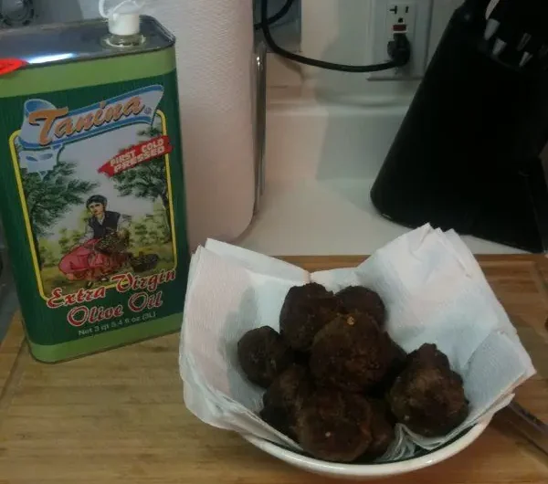 Bowl of homemade Italian meatballs resting on paper towels next to a tin of extra virgin olive oil on a kitchen counter.