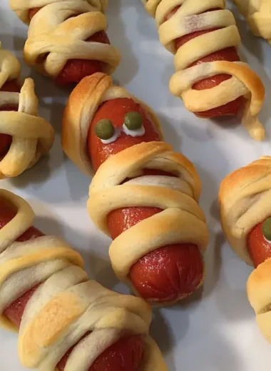 Finished mummy hot dogs with olive eyes wrapped in golden crescent roll strips on white parchment paper