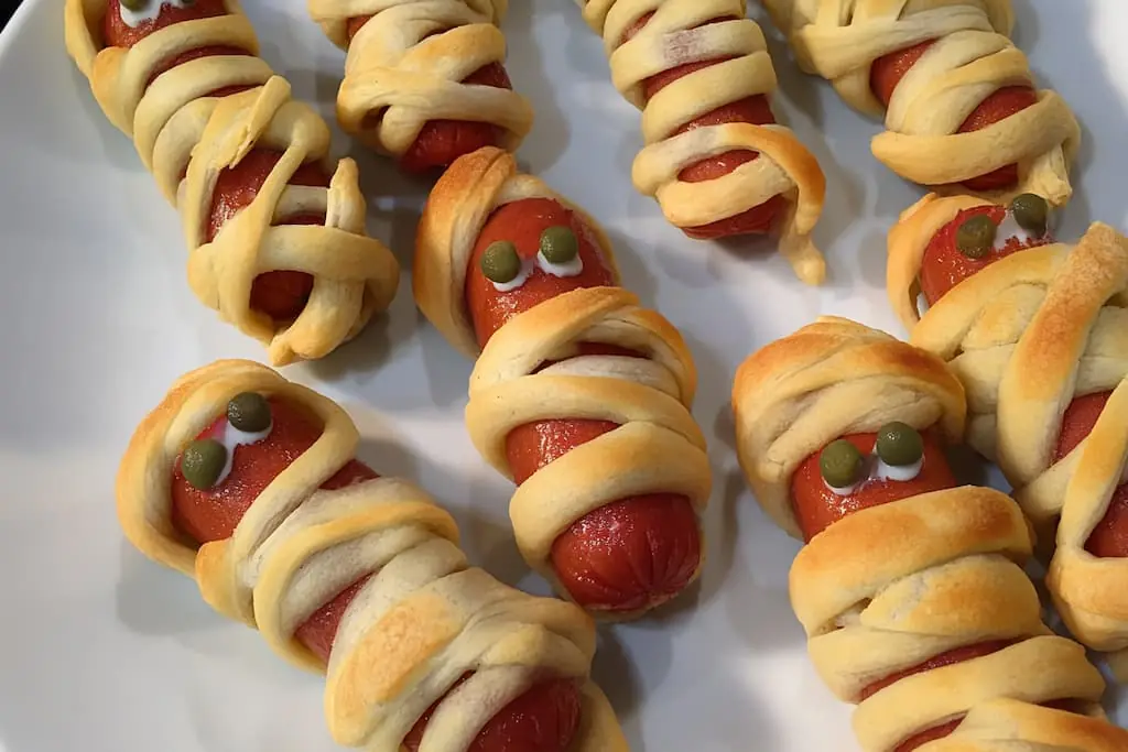 Finished mummy hot dogs with olive eyes wrapped in golden crescent roll strips on white parchment paper