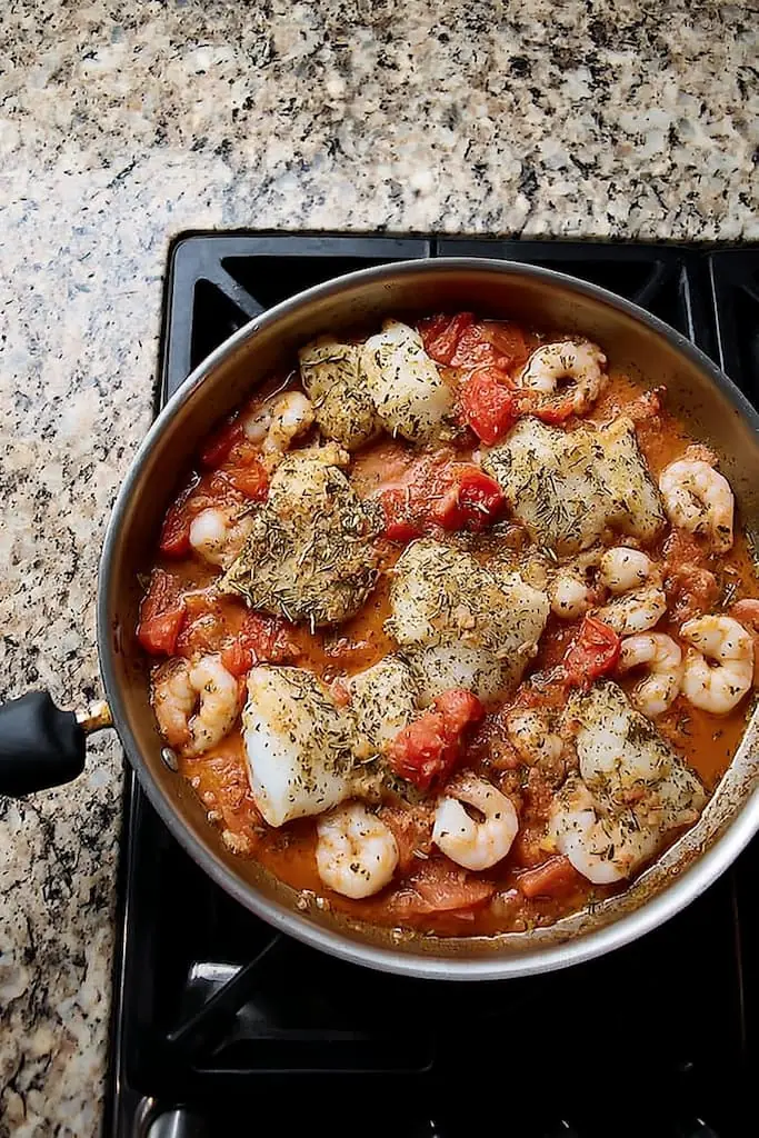 Overhead view of cod fillets and shrimp cooking in a stainless steel skillet with chunky tomato sauce, herbs, and seasonings on a gas stovetop