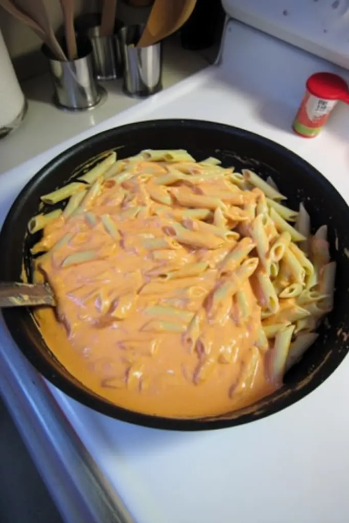 Penne pasta being tossed with creamy vodka sauce in a black non-stick pan, showing the pink sauce coating the noodles during the mixing process