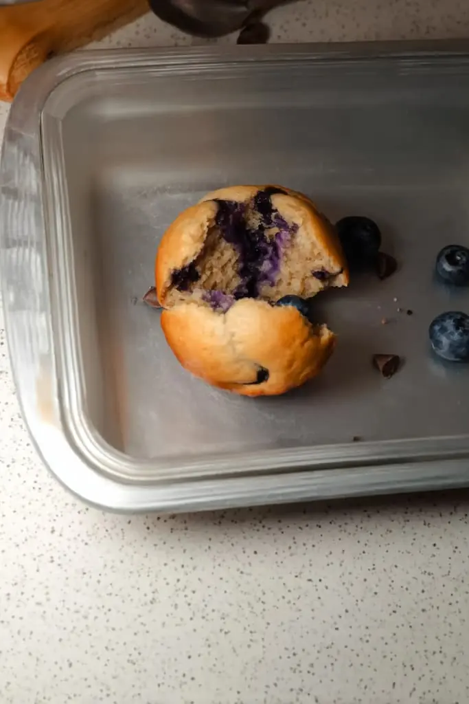 Split protein muffin showing fluffy interior crumb with bursted blueberries and chocolate chips in glass container