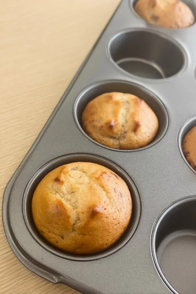 Freshly baked high protein banana muffins in a nonstick muffin tin showing perfectly risen golden-brown dome tops with natural banana specks visible in the batter