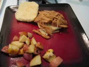 Pulled pork sandwich served on a bun with roasted red potatoes on a red plate.