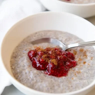 White ceramic bowl filled with creamy rose coconut chia seed pudding, topped with bright raspberry jam and a light cinnamon sprinkle, silver spoon resting inside.