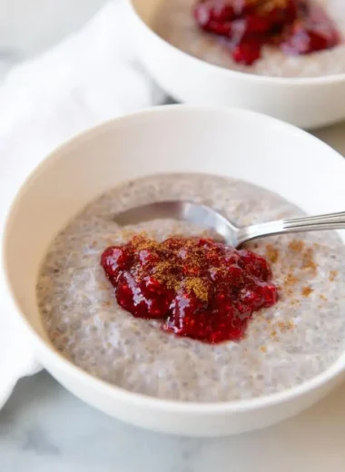 White ceramic bowl filled with creamy rose coconut chia seed pudding, topped with bright raspberry jam and a light cinnamon sprinkle, silver spoon resting inside.