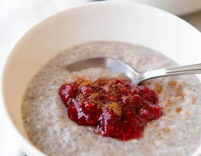 White ceramic bowl filled with creamy rose coconut chia seed pudding, topped with bright raspberry jam and a light cinnamon sprinkle, silver spoon resting inside.