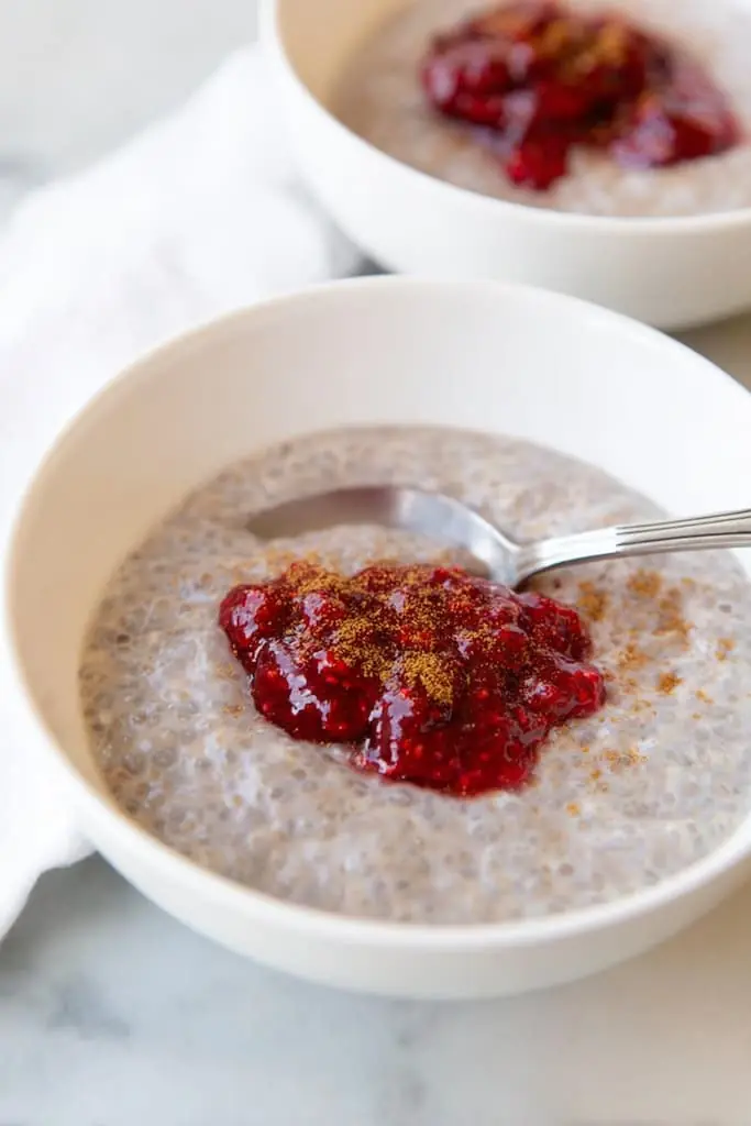 White ceramic bowl filled with creamy rose coconut chia seed pudding, topped with bright raspberry jam and a light cinnamon sprinkle, silver spoon resting inside.