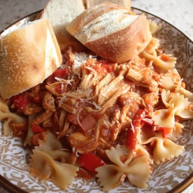 Plated slow cooker chicken cacciatore with shredded chicken, bell peppers, and onions over bow tie pasta, topped with Parmesan cheese and served with crusty Italian bread rolls