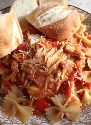 Plated slow cooker chicken cacciatore with shredded chicken, bell peppers, and onions over bow tie pasta, topped with Parmesan cheese and served with crusty Italian bread rolls