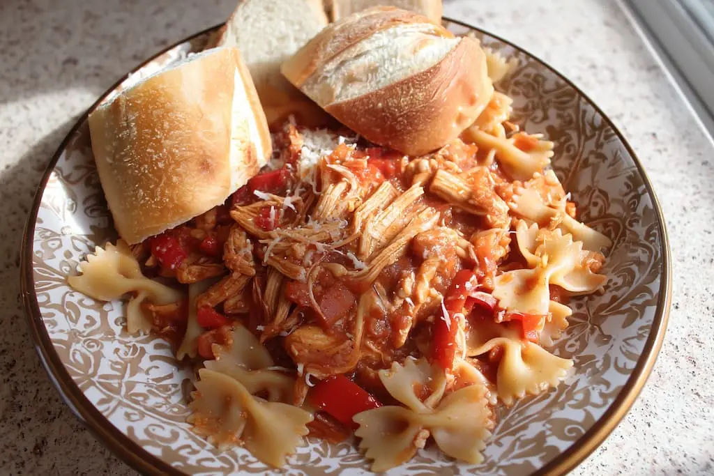 Plated slow cooker chicken cacciatore with shredded chicken, bell peppers, and onions over bow tie pasta, topped with Parmesan cheese and served with crusty Italian bread rolls