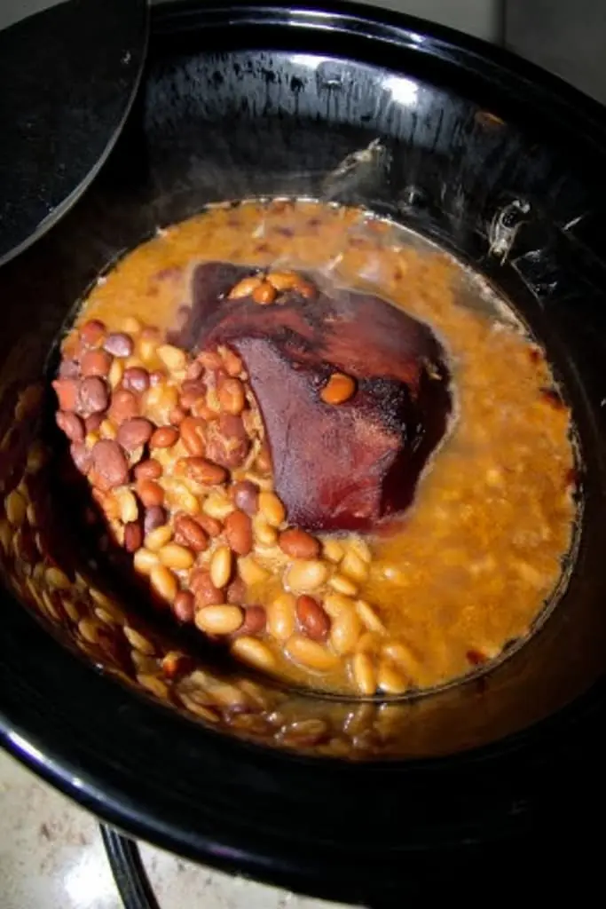 Soup beans with ham hock cooking in slow cooker showing mixed pinto and great northern beans with large ham hock in golden broth during cooking process