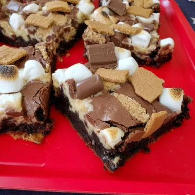 Gooey s'mores brownies topped with toasted mini marshmallows, melty milk-chocolate squares, and crunchy graham-cracker pieces on a red tray.