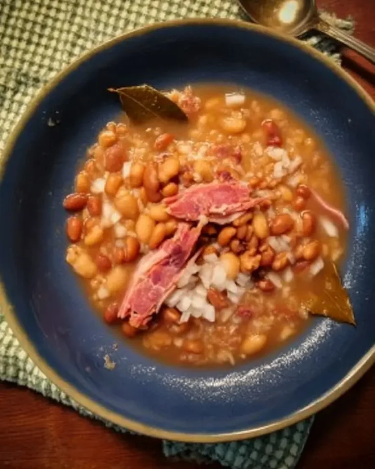 Bowl of soup beans with ham hock featuring tender pinto and great northern beans in rich broth with shredded ham pieces and chopped raw onions in blue ceramic bowl