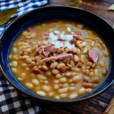 Soup beans with ham hock in dark blue ceramic bowl topped with chopped white onions and shredded ham pieces served on wooden table with checkered napkin