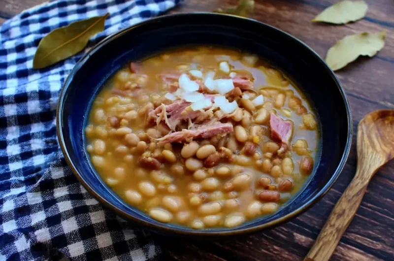 Soup beans with ham hock in dark blue ceramic bowl topped with chopped white onions and shredded ham pieces served on wooden table with checkered napkin