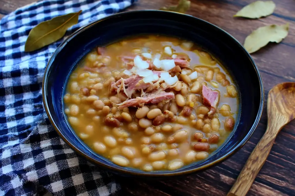 Soup beans with ham hock in dark blue ceramic bowl topped with chopped white onions and shredded ham pieces served on wooden table with checkered napkin