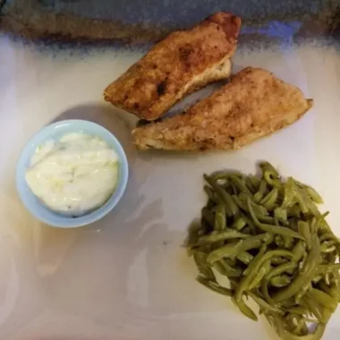 Square stoneware plate holding two golden fried catfish fillets, a scoop of tender green beans, and a small blue ramekin filled with creamy tartar sauce.
