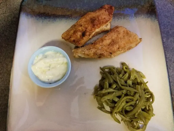 Square stoneware plate holding two golden fried catfish fillets, a scoop of tender green beans, and a small blue ramekin filled with creamy tartar sauce.