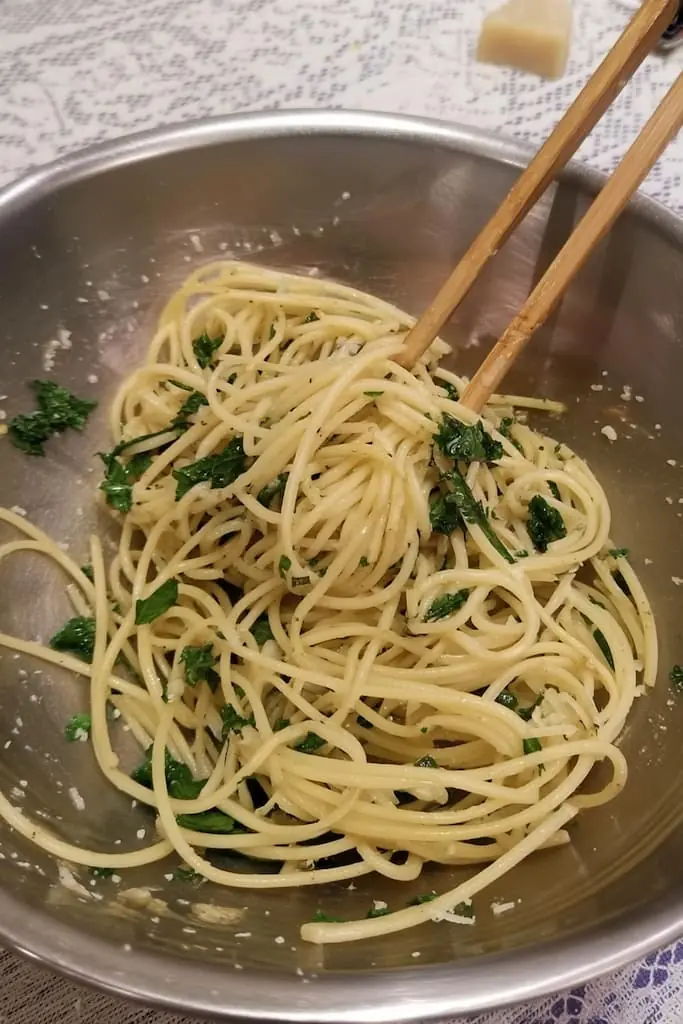 Spaghetti being tossed with wooden pasta forks in stainless steel bowl, showing pasta strands coated in golden garlic oil with fresh parsley and grated cheese